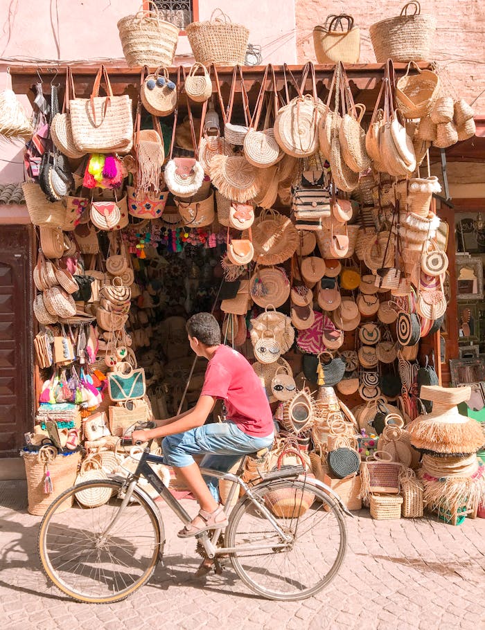 hero-img-02 A vibrant street market stall selling various woven bags with a person cycling by.